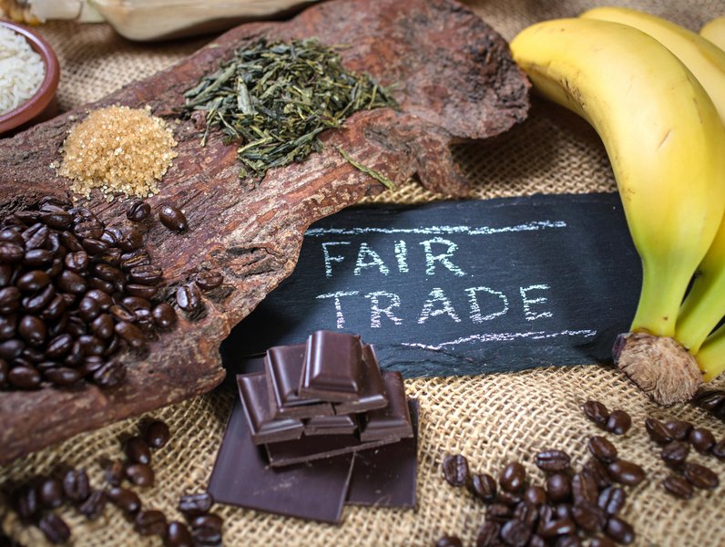 Kaffeebohnen, Zucker, Tee und Bananen liegen auf einem Stück Holz neben Schokoladenstücken. Eine Tafel mit der Aufschrift "FAIR TRADE" befindet sich auf einem groben Stoffuntergrund.