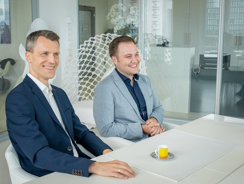Zwei Männer in Anzügen sitzen lächelnd an einem Tisch in einem modernen Büro mit Glastrennwänden. Zwischen ihnen steht eine gelbe Espressotasse auf einem Untersetzer.