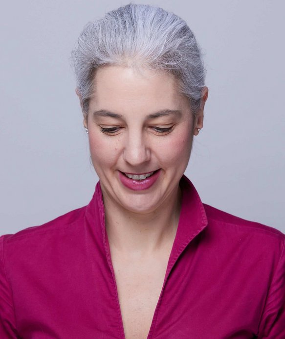 Frau mit grauem Haar lächelt und blickt nach unten. Sie trägt eine dunkelrosa Bluse. Der Hintergrund ist schlicht und grau.