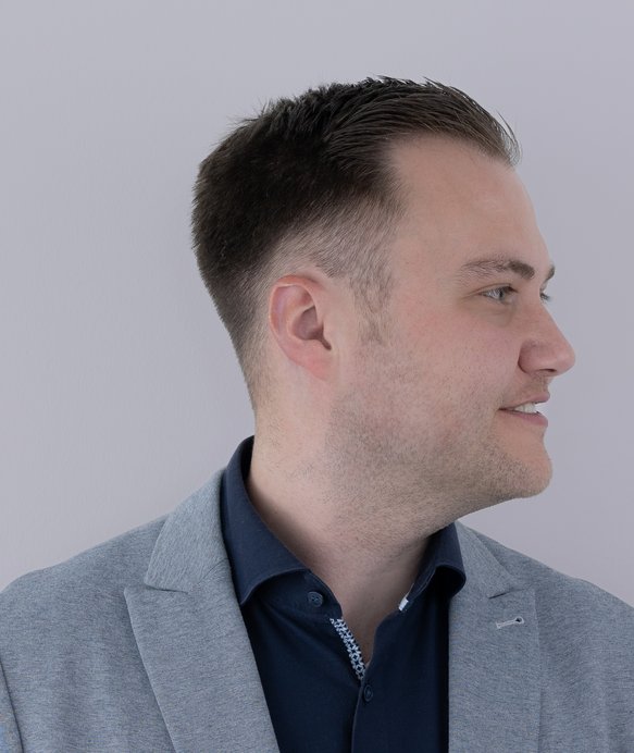 Ein Mann im Profil trägt ein graues Sakko über einem dunkelblauen Hemd, blickt nach rechts und steht vor einem hellgrauen Hintergrund.