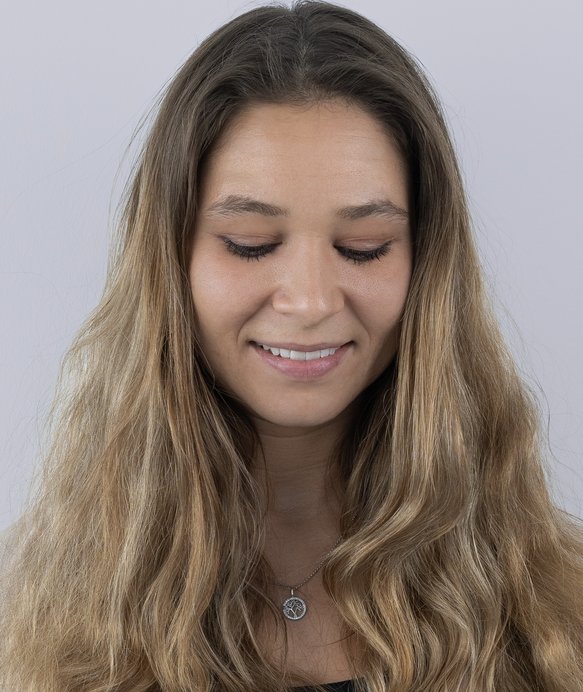 Eine Frau mit langen, welligen Haaren lächelt und schaut nach unten. Sie trägt eine Halskette mit Anhänger, vor einem neutralen, hellgrauen Hintergrund.