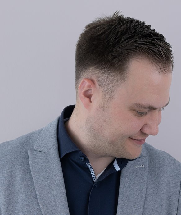 Ein Mann blickt leicht nach unten und links, trägt ein graues Sakko über einem dunkelblauen Hemd, vor einem neutralen, grauen Hintergrund.