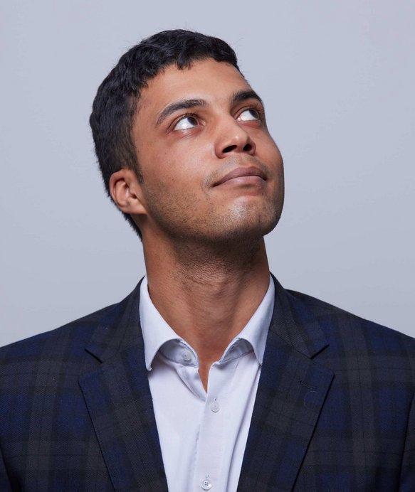 Ein Mann in einem karierten Anzug schaut nach oben. Der Hintergrund ist neutral grau. Sein Gesichtsausdruck wirkt nachdenklich. Keine weiteren Objekte oder Texte sind zu sehen.