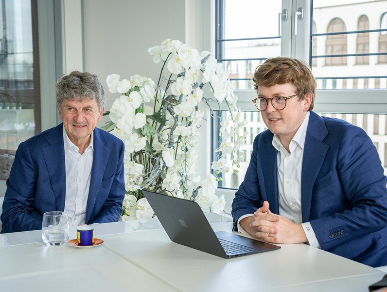 Zwei Männer in blauen Anzügen sitzen an einem Tisch. Einer bedient ein Laptop, während auf dem Tisch eine Tasse Kaffee und ein Wasserglas stehen. Im Hintergrund stehen weiße Blumen.