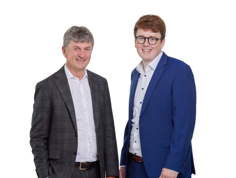 Zwei Männer in Business-Anzügen stehen nebeneinander, lächeln in die Kamera. Der linke Mann trägt einen grauen Anzug, der rechte einen blauen. Der Hintergrund ist schlicht weiß.