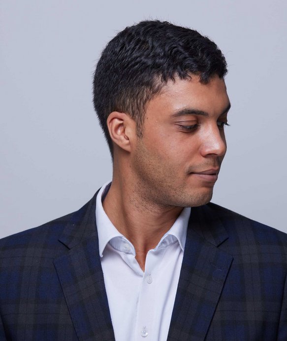 Ein Mann in einem karierten Sakko schaut nach unten. Der Hintergrund ist einfarbig grau, was den Fokus auf die Person verstärkt.