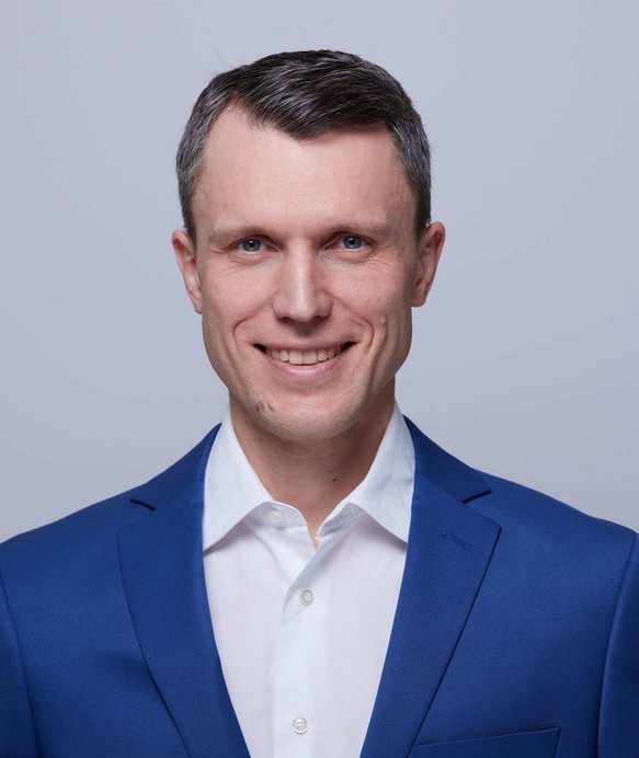 Ein Mann trägt einen blauen Anzug und ein weißes Hemd, lächelt direkt in die Kamera. Der Hintergrund ist schlicht grau, was den Fokus auf das Porträt lenkt.