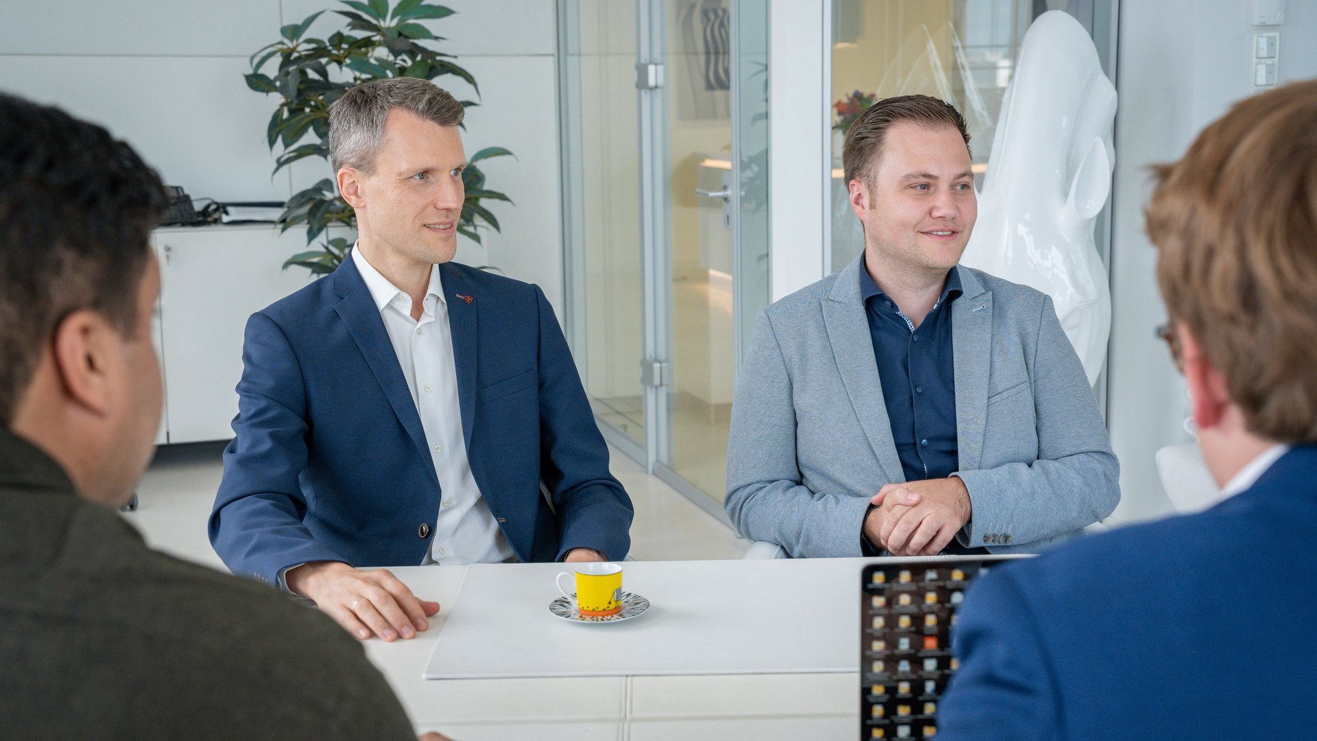 Zwei Männer in Anzügen sitzen an einem Tisch, unterhalten sich und schauen interessiert. Im Hintergrund moderne Büropflanzen und Glastüren. Zwei weitere Personen sind teilweise im Vordergrund zu sehen.