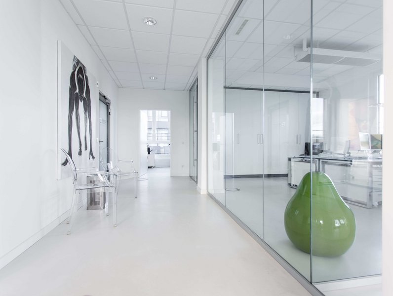 Transparente Stühle stehen in einem modern gestalteten, lichtdurchfluteten Flur mit Glastrennwand, während ein grüner, birnenförmiger Sitzsack in einem angrenzenden Büro mit weißen Möbeln ruht.
