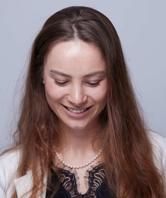 Eine Frau mit langen braunen Haaren lächelt sanft und schaut nach unten. Sie trägt eine Perlenkette und ein schwarzes Spitzentop. Der Hintergrund ist grau und schlicht.