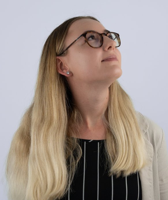 Eine Person mit schulterlangem, blondem Haar trägt eine Brille und schaut zur Seite. Sie trägt eine beigefarbene Jacke und ein schwarz gestreiftes Oberteil. Der Hintergrund ist neutral.