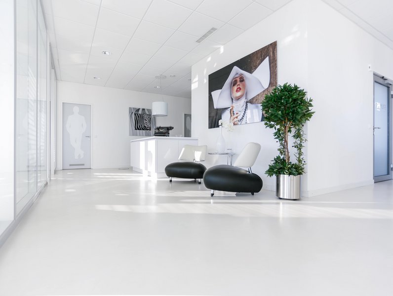 Zwei moderne schwarze Stühle stehen in einem hellen, minimalistisch eingerichteten Raum. Eine große Kunstfotografie hängt an der Wand, neben einer grünen Topfpflanze. Links ist eine Glaswand sichtbar.