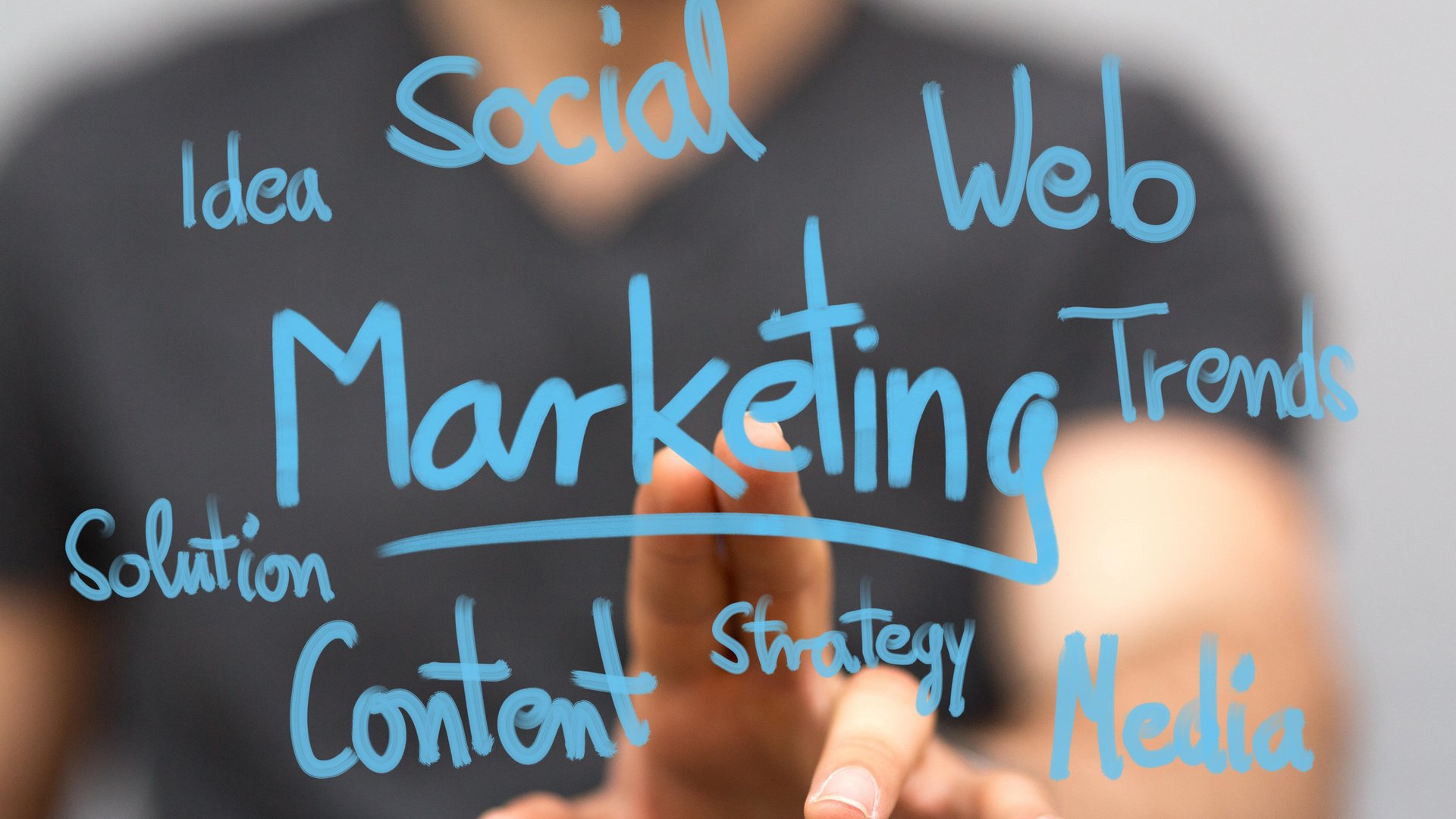 Eine Hand zeigt auf Wörter wie "Marketing," "Social," "Web," "Content," und "Trends," die in blauer Schrift auf Glas geschrieben sind. Im Hintergrund ist eine unscharfe Person.