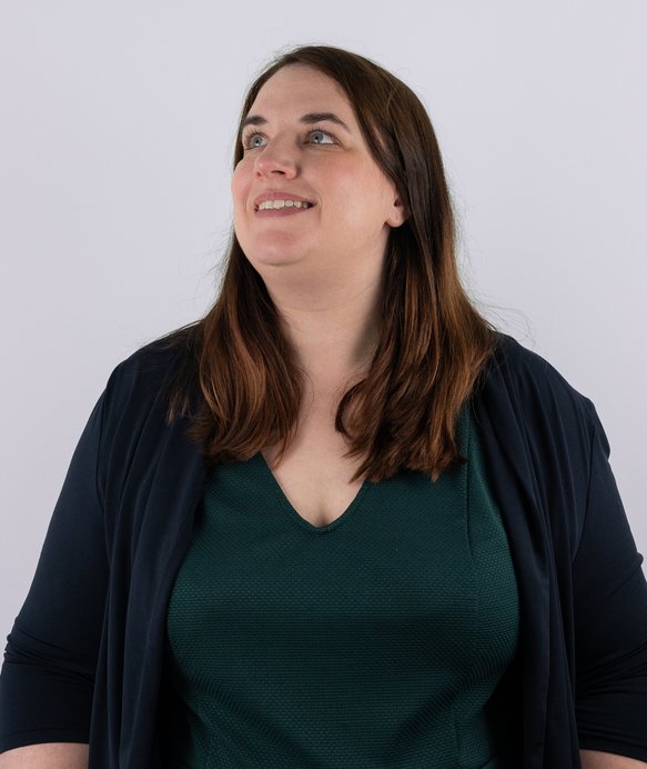 Eine Frau mit schulterlangem, braunem Haar blickt nach oben und lächelt. Sie trägt ein dunkles Oberteil und steht vor einem neutralen, weißen Hintergrund.