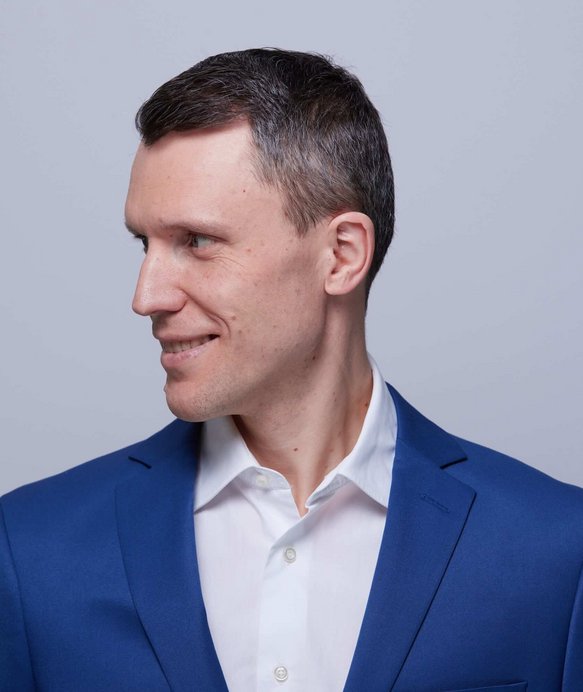 Ein Mann in einem blauen Anzug blickt lächelnd zur Seite, vor einem grauen Hintergrund. Sein Haar ist kurz geschnitten, und er trägt ein weißes Hemd unter dem Anzug.