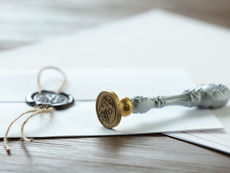 Ein goldener Siegelstempel liegt auf einem weißen Umschlag. Der Umschlag ist mit einem verschnürten Wachssiegel verschlossen. Der Hintergrund zeigt eine Holzoberfläche.