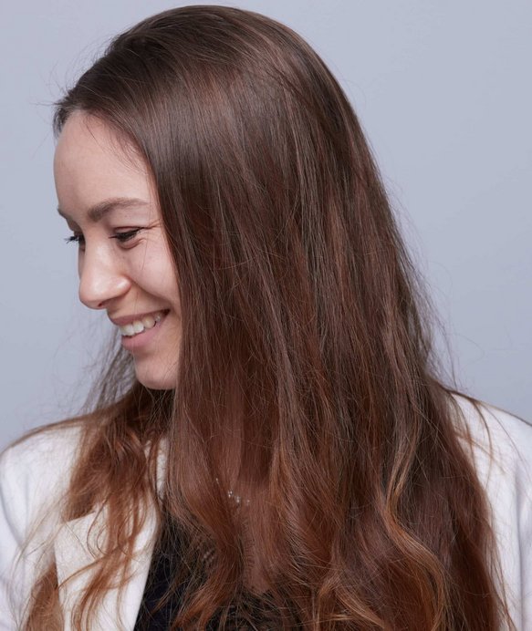 Eine Frau mit langen, glatten Haaren lächelt und blickt leicht nach unten. Sie trägt ein weißes Oberteil. Der Hintergrund ist einfarbig grau.