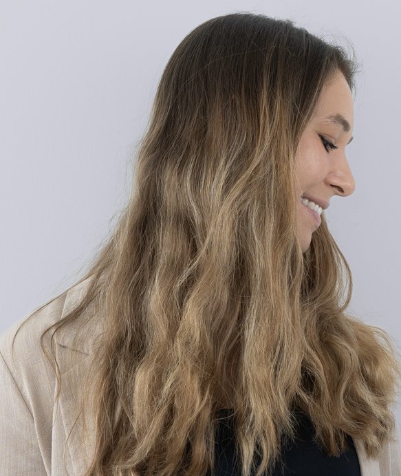 Frau mit langen, welligen Haaren lächelt, während sie den Kopf zur Seite neigt. Sie trägt eine beige Jacke und das Bild hat einen neutralen, hellen Hintergrund.