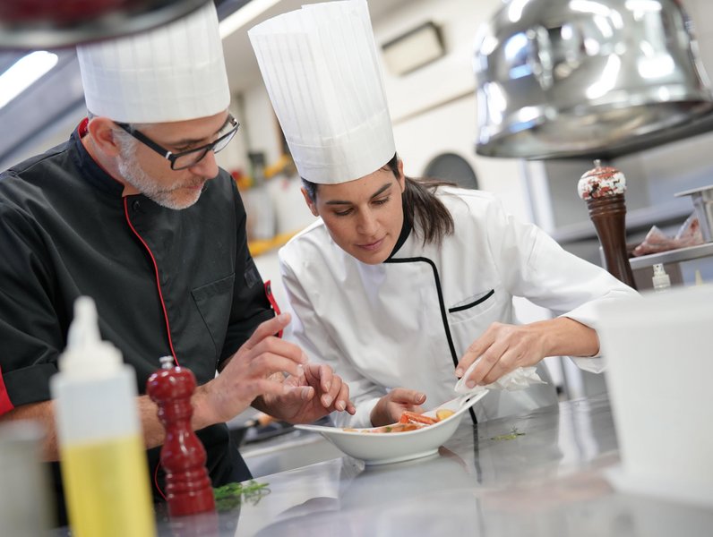 Zwei Köche tragen weiße Mützen und richten sorgfältig ein Tellergericht an. Sie befinden sich in einer professionellen Restaurantküche mit Utensilien und Gewürzen im Hintergrund.