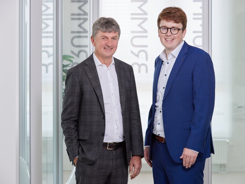 Zwei Männer stehen nebeneinander und lächeln in die Kamera. Sie tragen Anzüge; der ältere Mann einen dunklen, der jüngere einen blauen. Im Hintergrund sind Glaswände mit Schrift.