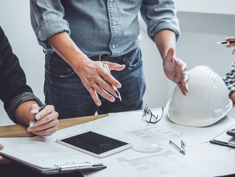 Personen diskutieren mit gestikulierenden Händen über Baupläne, umgeben von einem Tablet, einem Bauhelm, einer Brille und Stiften auf einem Tisch in einem Büro.