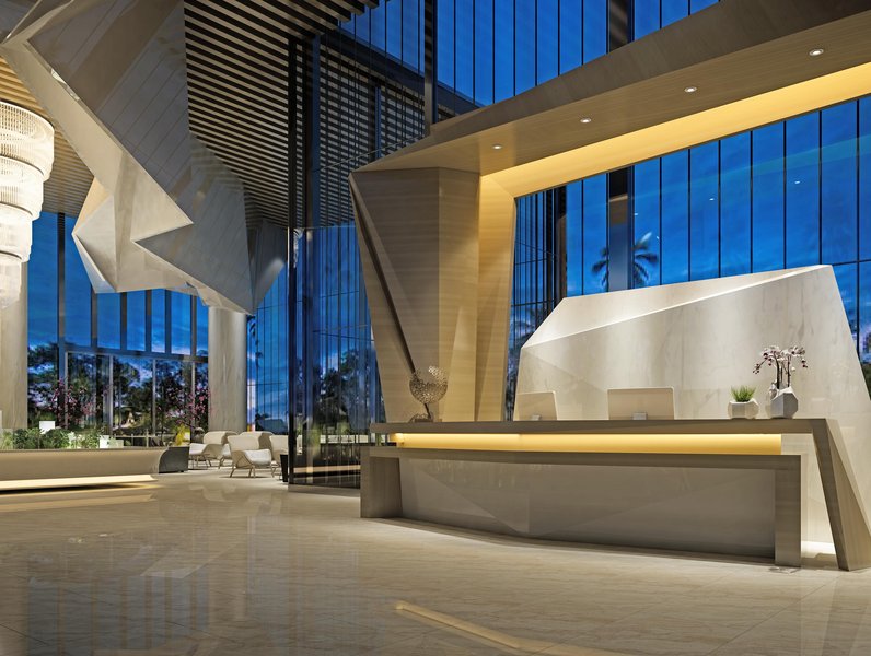 Empfangstheke aus Marmor steht beleuchtet in einem modernen, geräumigen Foyer mit hohen Decken und großen Glasfenstern. Dekorative Pflanzen und stilvolle Sitzmöbel ergänzen die elegante Atmosphäre.