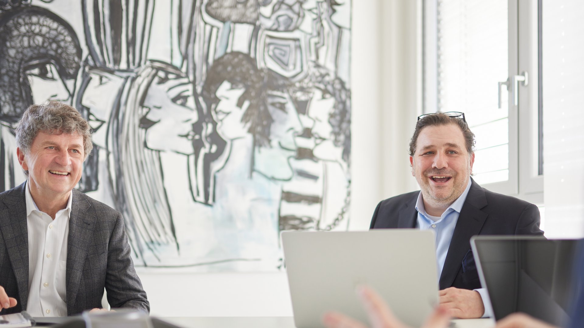 Zwei Männer lächeln, während sie an einem Tisch mit Laptops sitzen, vor einem großen, abstrakten Wandbild in einem hellen Büro mit Fenstern.