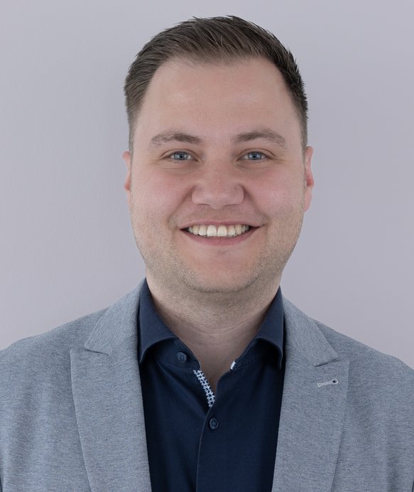 Mann lächelt freundlich, trägt ein hellgraues Sakko und ein dunkles Hemd, steht vor einem neutralen, hellgrauen Hintergrund.