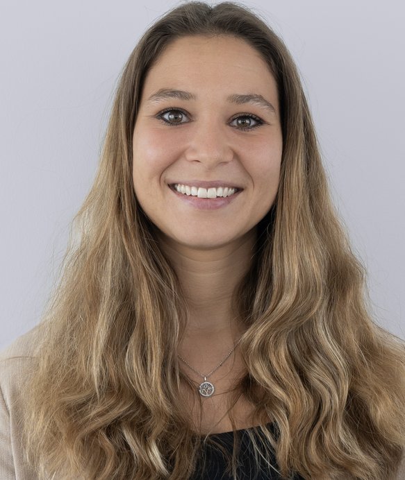 Eine Frau mit langen, welligen Haaren lächelt in die Kamera. Sie trägt eine Halskette mit Anhänger und steht vor einem hellgrauen Hintergrund.