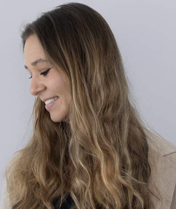 Frau mit langen, braunen Haaren lächelt und blickt nach unten. Sie trägt eine helle Jacke und steht vor einem neutralen, hellgrauen Hintergrund.