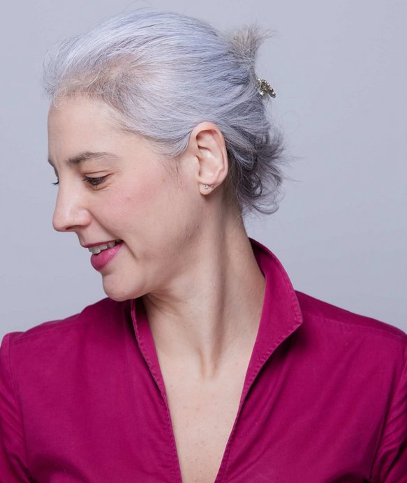 Frau mit grauem Haar sieht zur Seite. Sie trägt eine pinke Bluse, Haare zurückgesteckt. Der Hintergrund ist neutral grau.