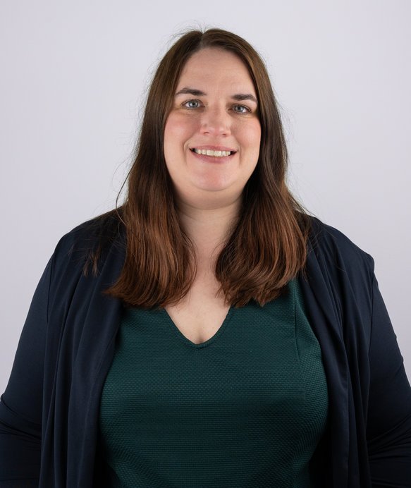 Eine Frau lächelt direkt in die Kamera, trägt ein dunkelgrünes Oberteil und einen dunkelblauen Schal. Sie steht vor einem schlichten, weißen Hintergrund. Keine weitere Umgebung erkennbar.