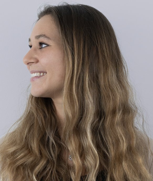 Eine Frau mit welligem, braunem Haar lächelt und blickt nach links vor einem neutralen, hellgrauen Hintergrund.