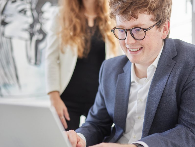 Ein lächelnder Mann arbeitet am Laptop, während eine Frau im Hintergrund steht. Sie befinden sich in einem hellen Büro mit einer modernen Zeichnung an der Wand.
