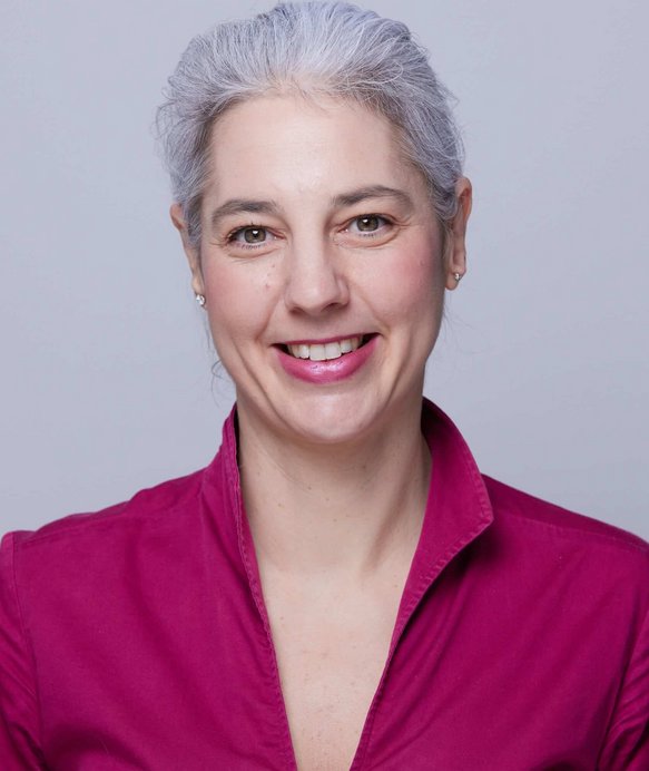 Eine lächelnde Frau mit grauem Haar trägt ein pinkfarbenes Hemd. Sie steht vor einem schlichten, hellgrauen Hintergrund.