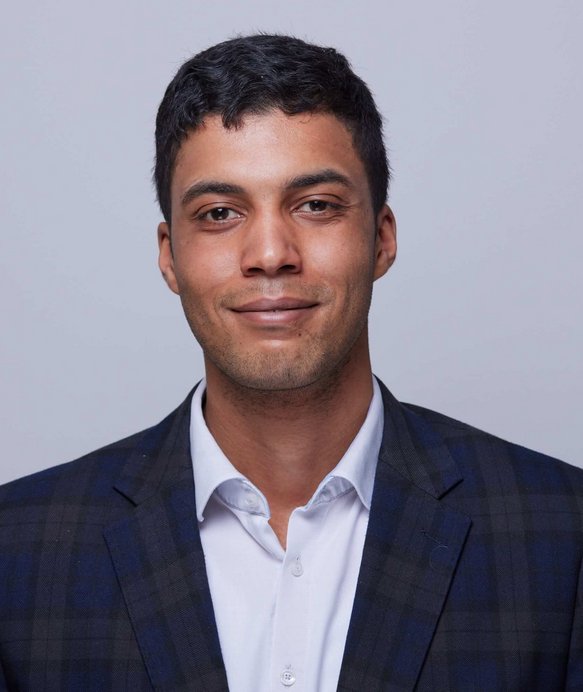 Ein Mann trägt einen dunklen Karoblazer und ein weißes Hemd und blickt lächelnd in die Kamera. Der Hintergrund ist neutral und hell, wodurch er im Fokus steht.