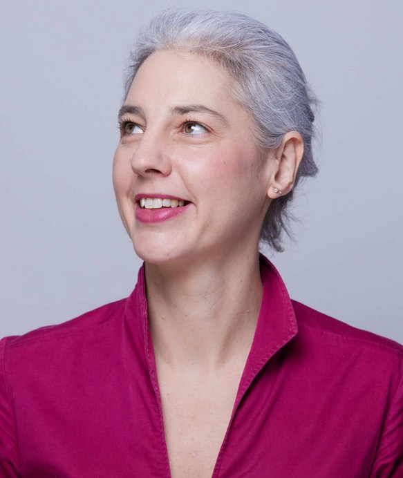 Eine Frau mit grauem Haar lächelt und schaut nach links oben. Sie trägt eine magentafarbene Bluse vor einem grauen Hintergrund.