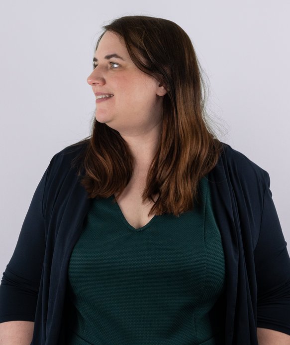 Eine Frau mit schulterlangem braunem Haar, trägt ein dunkelgrünes Oberteil und eine dunkelblaue Strickjacke, schaut lächelnd nach links vor einem neutralen, hellgrauen Hintergrund.