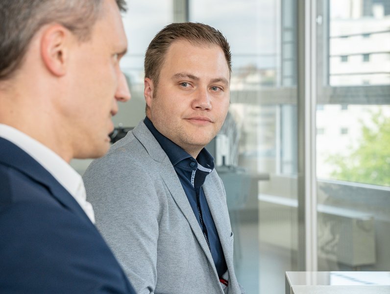 Ein Mann in grauem Anzug sitzt in einem modernen Büro und schaut seinen Gesprächspartner an. Im Hintergrund sind große Fenster und verschwommene Gebäude sichtbar.