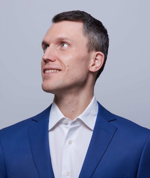 Ein Mann im blauen Anzug schaut lächelnd nach links oben, vor einem einfarbigen, hellgrauen Hintergrund. Er trägt ein weißes Hemd unter dem Anzug.