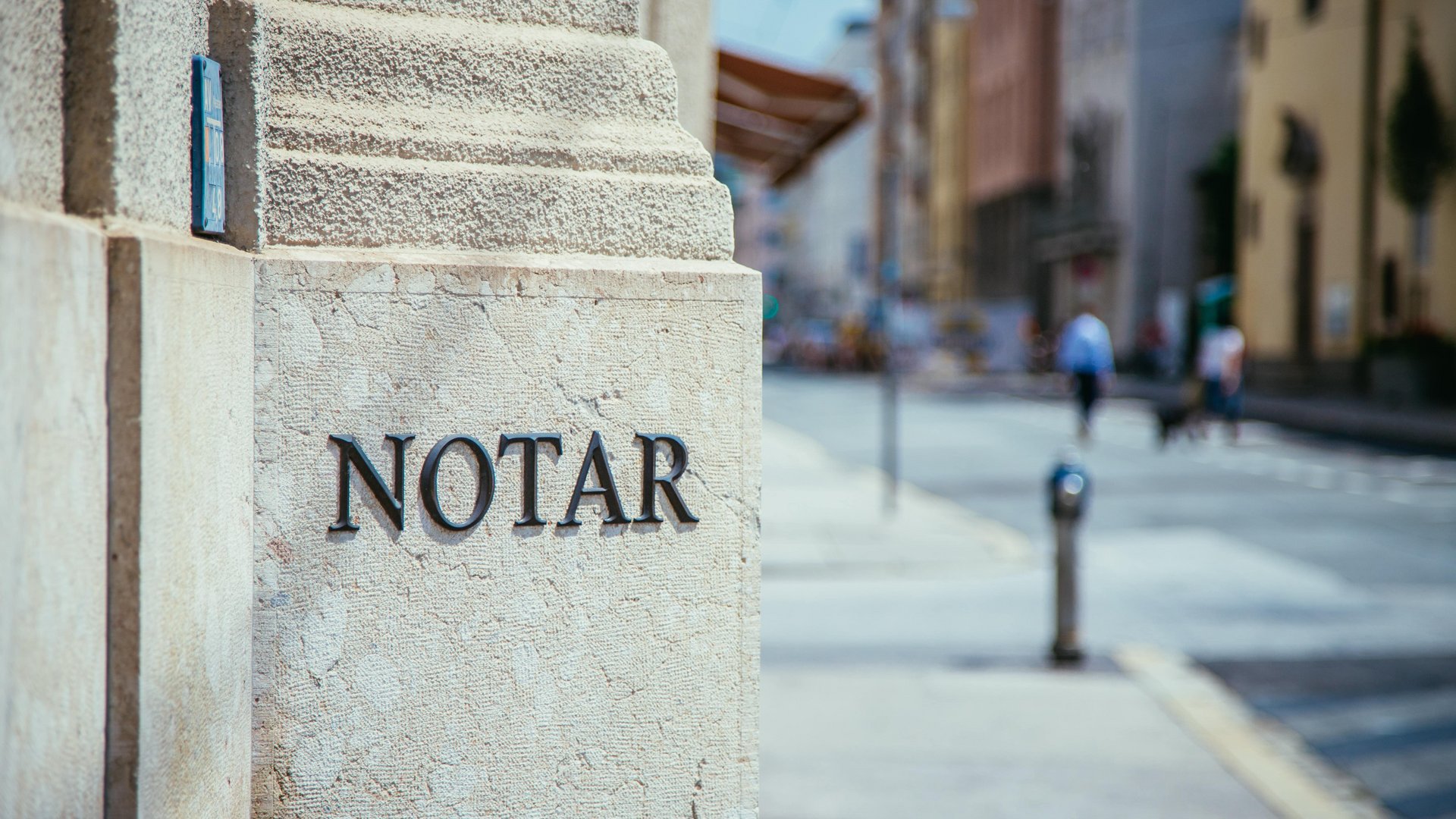 Steinwand mit schwarzem Schriftzug "NOTAR", im Hintergrund eine unscharfe Straßenszene mit Gehenden und Gebäuden.