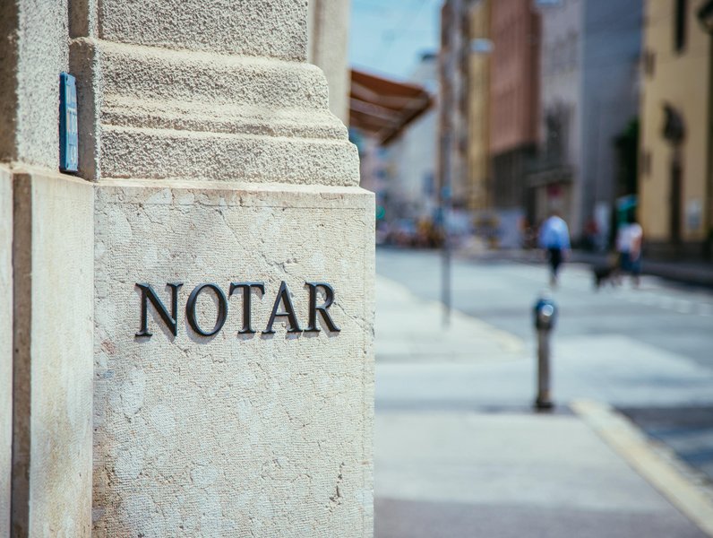 Steinwand mit schwarzem Schriftzug "NOTAR", im Hintergrund eine unscharfe Straßenszene mit Gehenden und Gebäuden.