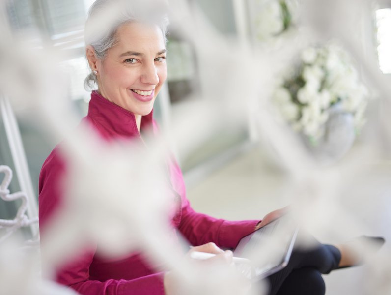 Frau in rotem Oberteil hält ein Tablet und lächelt. Sie sitzt entspannt in einem hellen Raum, umgeben von unscharfen, dekorativen Elementen. Im Hintergrund befinden sich Blumentöpfe.