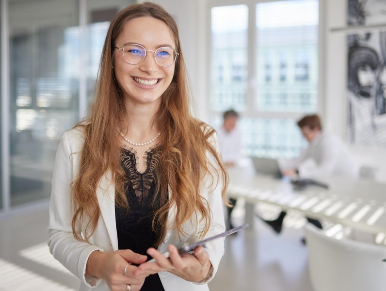 Eine Frau lächelt, hält ein Tablet in einem modernen Büro. Im Hintergrund arbeiten zwei Personen an Laptops an einem weißen Tisch. Die Umgebung ist hell und geräumig.