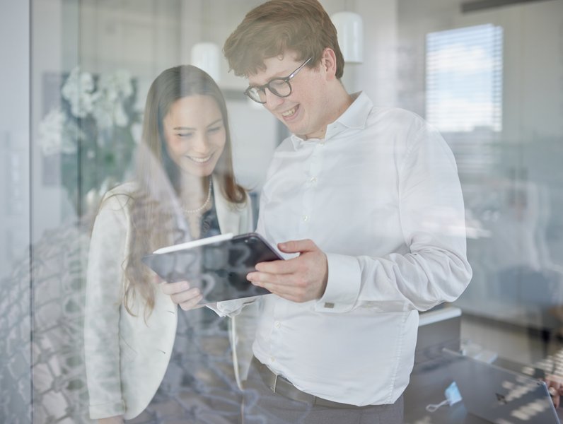 Zwei Personen betrachten gemeinsam ein Tablet, lächelnd und in Business-Kleidung. Sie stehen in einem modernen Büro mit Glaswänden und Lampen im Hintergrund.