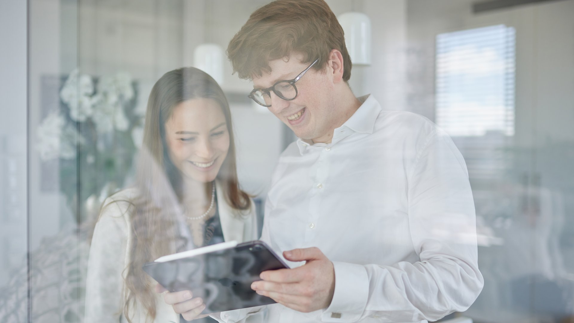 Zwei Personen betrachten gemeinsam ein Tablet, lächelnd und in Business-Kleidung. Sie stehen in einem modernen Büro mit Glaswänden und Lampen im Hintergrund.