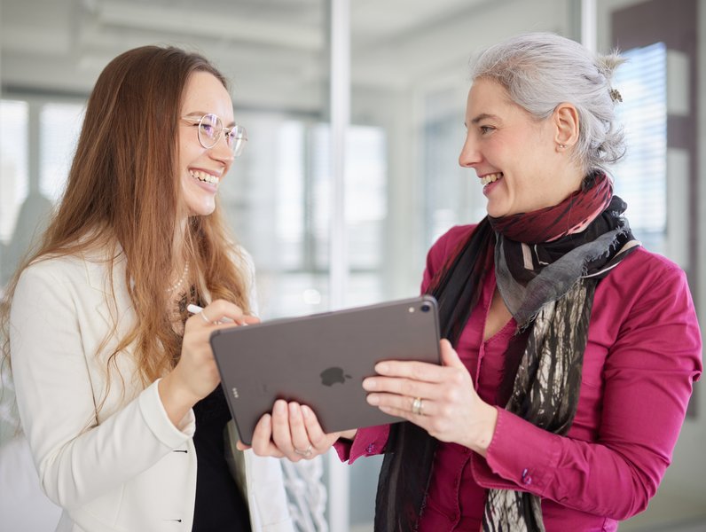 Zwei Frauen, halten ein Tablet und lächeln einander an. Sie stehen in einem modernen, lichtdurchfluteten Büro mit Glaswänden.
