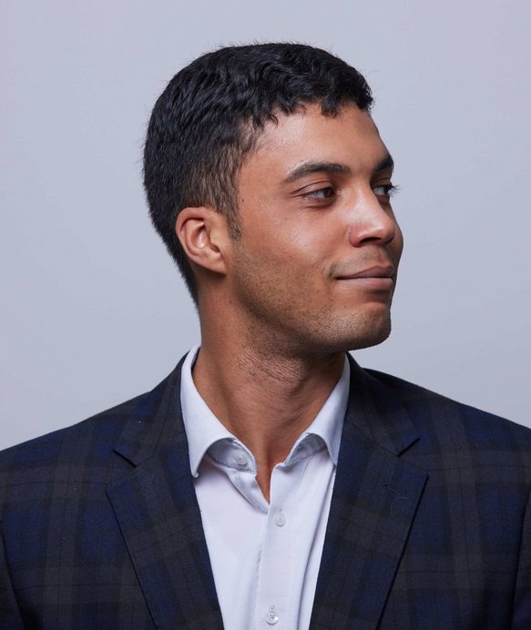 Ein Mann in einem karierten Anzug schaut lächelnd zur Seite. Der Hintergrund ist einfarbig hellgrau, was die Aufmerksamkeit auf sein Gesicht und seine Kleidung lenkt.