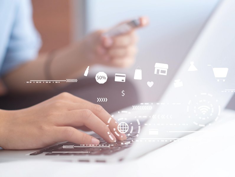 Hände tippen auf einer Laptop-Tastatur, während eine Person ein Smartphone hält. Schwebende Symbole und Icons, wie Einkaufswagen und Prozentsätze, verdeutlichen Online-Shopping in einem modernen, digitalen Umfeld.