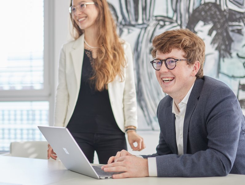 Ein Mann sitzt lächelnd am Laptop, während eine Frau neben ihm steht und ebenfalls lächelt. Beide befinden sich in einem modernen Büro mit kunstvollen Wandgemälden.